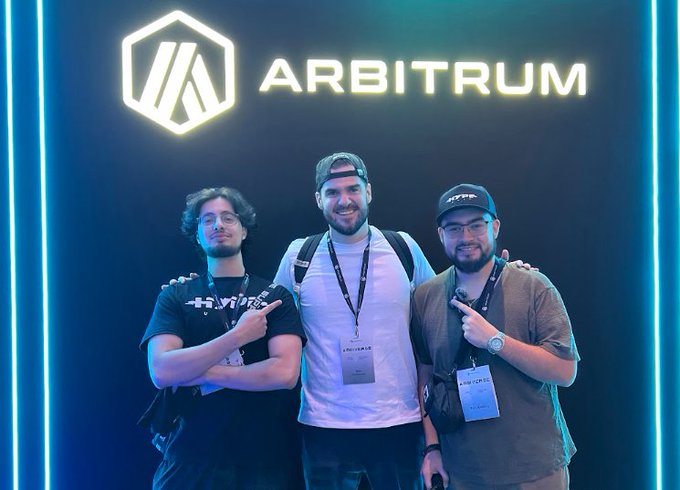 Three men pose together, smiling and pointing at a neon-lit 'ARBITRUM' logo on a dark blue wall.