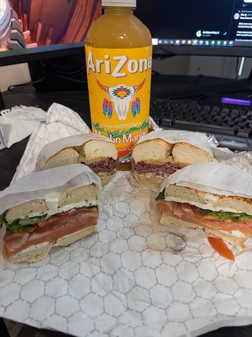 A simple "Gm" (good morning) setup featuring two halves of bagel sandwiches—one with smoked salmon and the other with pastrami—paired with an Arizona "Mucho Mango" juice bottle on a desk. The casual breakfast-on-the-go captures a relaxed morning routine before starting the workday.