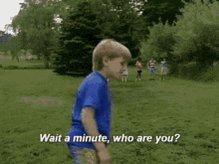 The image features a young boy wearing a blue shirt, standing in a grassy field with trees in the background. The text "WAIT A MINUTE, WHO ARE YOU?" is written above him, indicating that he has been chosen as the subject of an advertisement or promotion.
