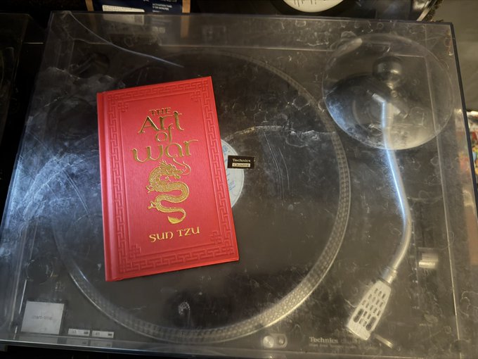 A small red leather-bound edition of "The Art of War" by Sun Tzu resting on the dust cover of a Technics turntable.