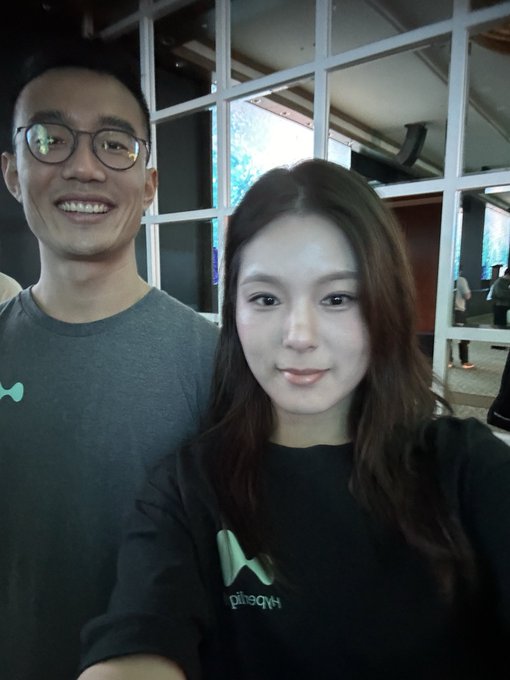 A man with glasses and a woman wearing a black "Hyperliquid" sweatshirt pose together for a selfie in what appears to be a modern indoor venue with large windows. The man wears a gray shirt with a small green logo, and they both smile at the camera, capturing a moment connected to the Hyperliquid community as mentioned in the post.