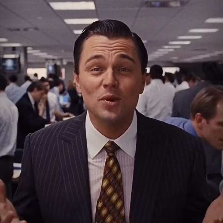 The image features a man dressed in a suit standing in an office setting with other people around him. The main subject of the photo appears to be a man wearing a suit, possibly a movie star or a businessman, who seems to be giving a speech or making a statement. There are multiple people present in the scene, including those sitting at desks and others standing nearby.

The office environment is filled with various objects such as chairs, TVs mounted on the walls, and other items like laptops and keyboards. The presence of these objects suggests that this setting could be an office or a workspace where people gather to work or discuss business matters.