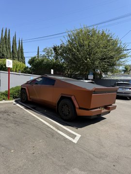 A matte orange/brown Tesla Cybertruck is parked in a parking lot under a clear blue sky.