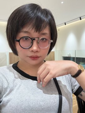 A selfie of a young person with short dark hair wearing glasses with pink-tinted lenses and a gray sweater, posing with their hand near their face while wearing a watch.