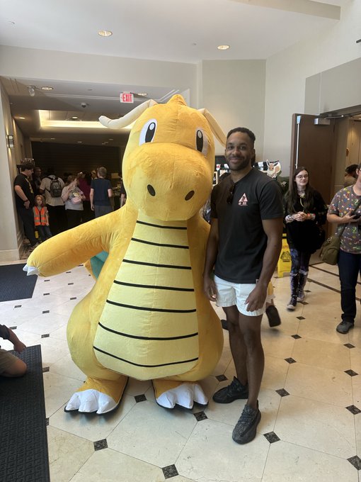 A man in a black t-shirt and white shorts poses next to a person wearing a Dragonite Pokémon mascot costume indoors.