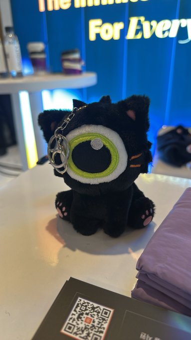 A black "ZKat" plush keychain with a large green eye sits on a white counter, with a QR code labeled "fly..." visible in the foreground and "The Future For Every" illuminated in the background, supporting the post about verifying with Brevis at KBW.