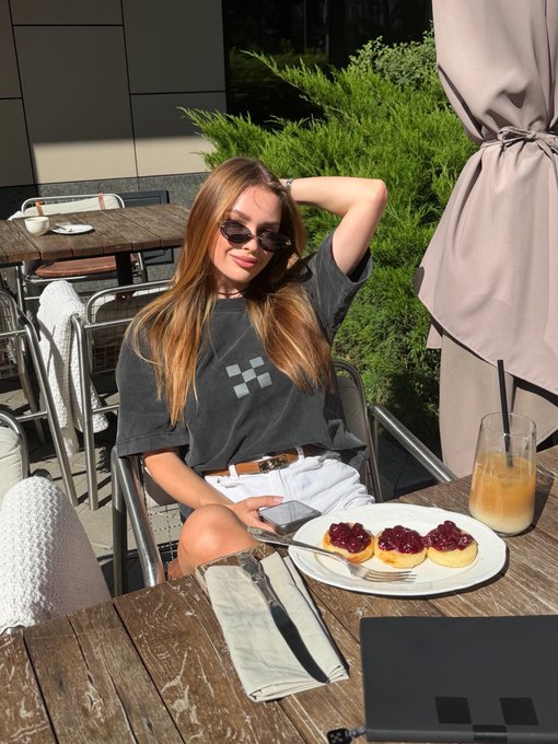 A young woman with long hair and sunglasses sits at an outdoor restaurant table wearing a black t-shirt, with a plate of jam-topped pastries and an orange drink in front of her.