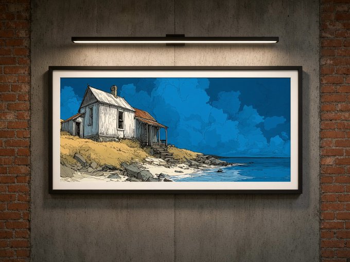 A framed illustration of a weathered coastal cottage on a beach under a dramatic blue sky, mounted on a concrete wall illuminated by a sleek black light fixture.