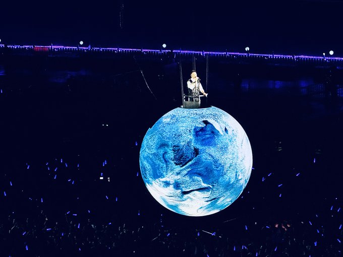 A performer sits on top of a large illuminated globe hanging above a dark concert arena filled with blue light sticks.