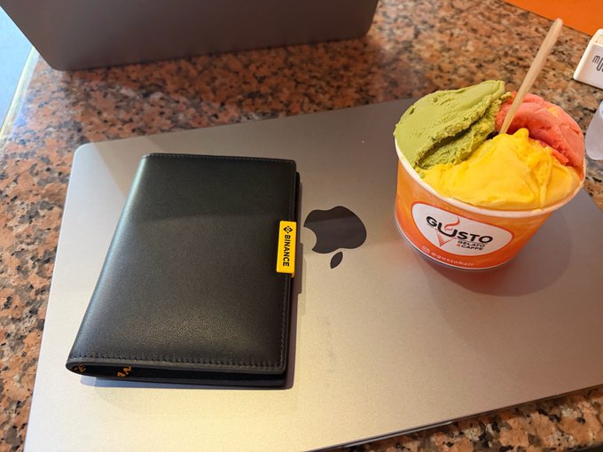 A black wallet with a yellow 'BINANCE' tag sits next to a cup of three-flavor gelato (green, yellow, and red/pink) from 'Giusto Gelato' on a MacBook laptop.