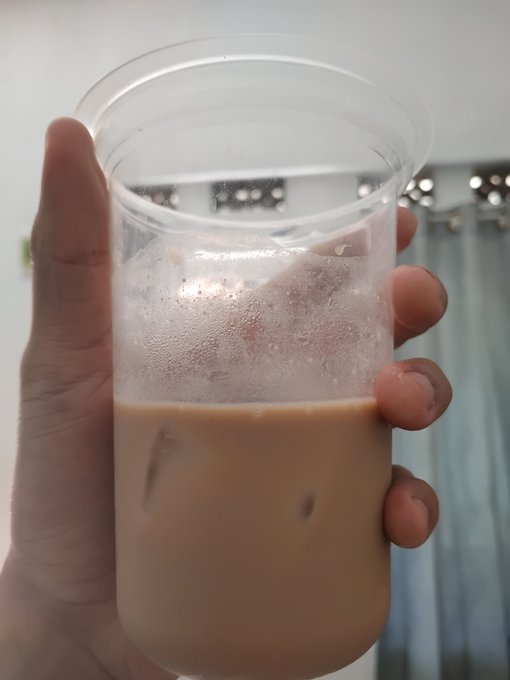 A hand holds up a clear plastic cup containing a milky iced coffee or milk tea, taken from an indoor location with ceiling lights visible in the background.