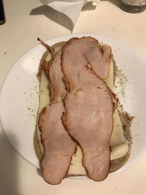 An open-faced sandwich on a white paper plate, layered with cheese and topped with multiple overlapping slices of ham, seasoned with pepper and herbs on a countertop.