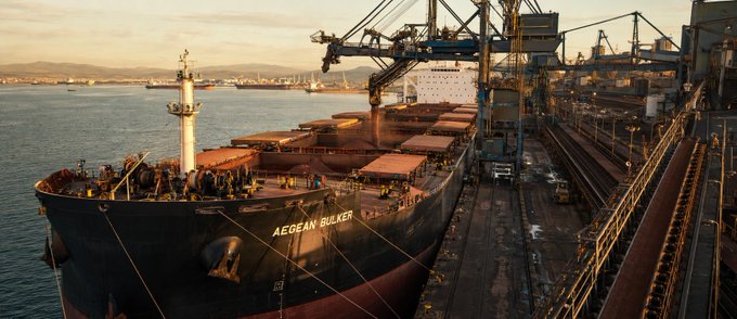 A bulk carrier ship named 'AEGEAN BULKER' is docked at an industrial port, where large cranes are loading reddish-brown bulk material (likely ore) into its cargo holds at golden hour.