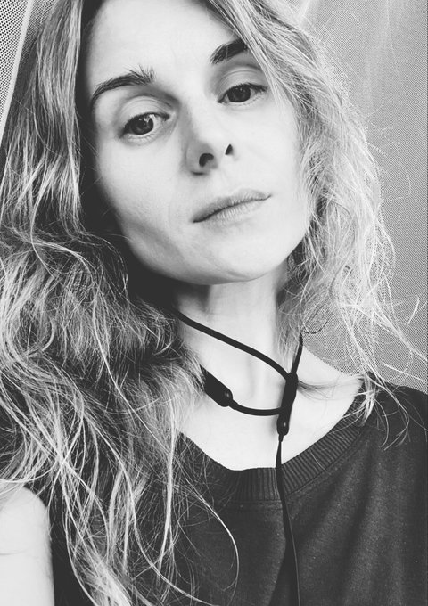 A black-and-white selfie portrait of a woman with long wavy hair, wearing a dark top and a black cord around her neck, looking directly at the camera.