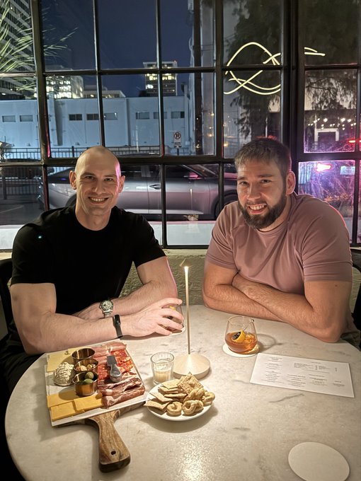 Two men sit smiling at a restaurant table at night, with a charcuterie board featuring meats and cheeses, a candle, drinks, and a menu visible in front of them.