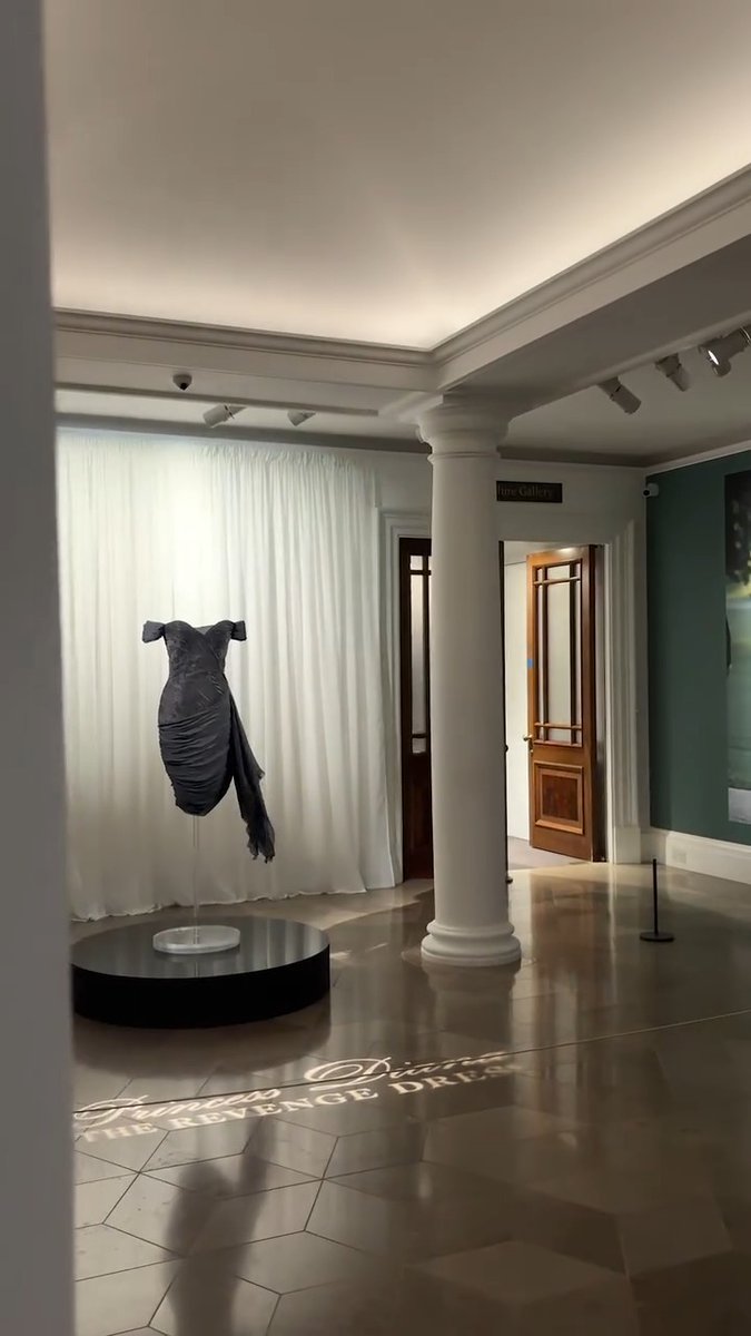 A museum gallery interior featuring an art installation with a dress-like sculpture displayed on a black circular platform, with text on the floor referencing 'Tom Ford' and 'The Revenge Dress'.