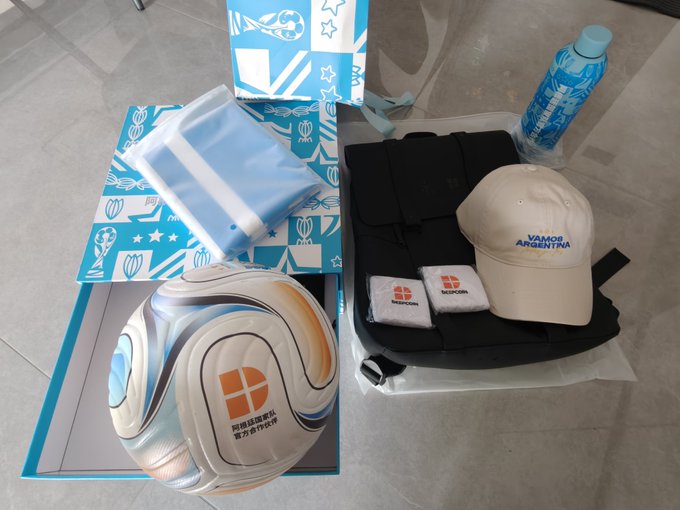 A photograph of promotional merchandise including a volleyball, blue and white patterned boxes, a beige 'VAMOS ARGENTINA' cap, black bags, wristbands, and a water bottle.