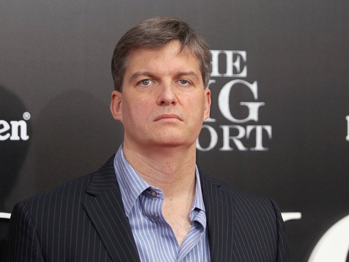 A man in a pinstripe suit and blue striped shirt poses in front of a backdrop with partially obscured text.