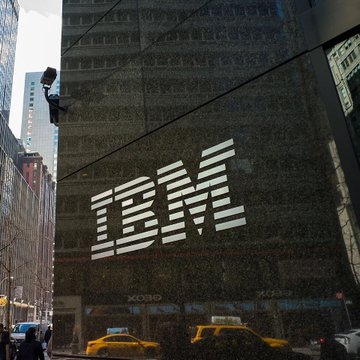 The image shows a tall building with the IBM logo prominently displayed on its side. The text "CRAZY" is also visible in the image. In front of the building, there are two yellow taxis driving by, one closer to the camera and another further away. Additionally, several people can be seen walking along the sidewalk near the building.