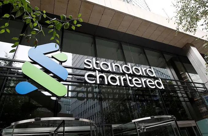 The image shows the exterior of a Standard Chartered bank building, featuring its distinctive blue and green logo alongside the "Standard Chartered" name displayed prominently on the glass facade. This visual directly complements the post's announcement, as it represents the global banking giant expanding its services to include spot Bitcoin and Ether trading for institutional clients in the UAE.