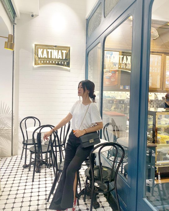 A woman in a white t-shirt and black slit skirt sits on a black bentwood chair outside a 'KATINAT' café, with a tiled black-and-white floor and a glass display case visible behind her.