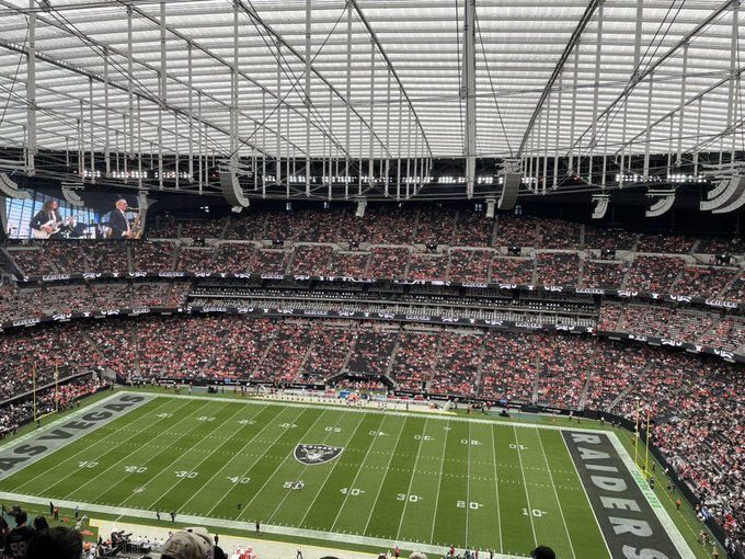A wide view of a packed Las Vegas Raiders football stadium with the field logo visible at midfield and a large screen showing people in the stands.