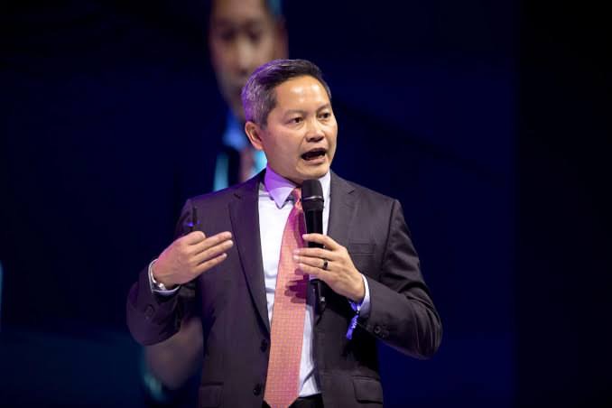 A man in a dark suit with a pink tie speaking into a microphone while gesturing, standing in front of a large screen displaying his face.