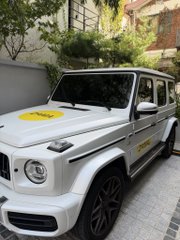 A white Mercedes-Benz G-Class SUV parked outdoors with a yellow promotional sign on the hood.