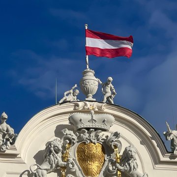 An ornate baroque building facade is topped with an Austrian flag (red-white-red) waving against a clear blue sky, featuring a golden coat of arms and classical sculptural figures. The image visually anchors the Austria-focused news about the FMA's first MiCA penalty against Bitpanda, with the national flag symbolizing the Austrian regulatory authority behind the €70,000 fine.