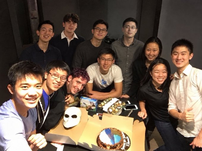 A group photo of about ten young people gathered around a table holding a white mask, a tiered cake, and a box of donuts, seemingly at a celebration.