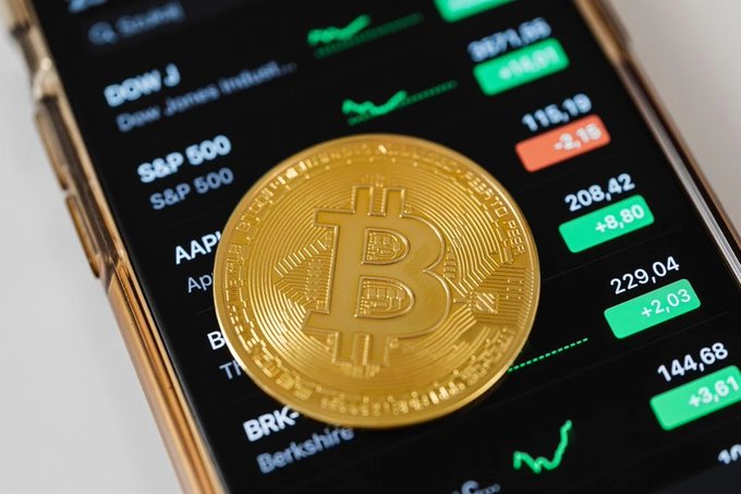 A close-up photograph of a physical Bitcoin coin resting on a smartphone screen displaying a stock market app with various tickers including DOW J (Dow Jones Indus), S&P 500, and AAPL, with green positive indicators and red negative ones.