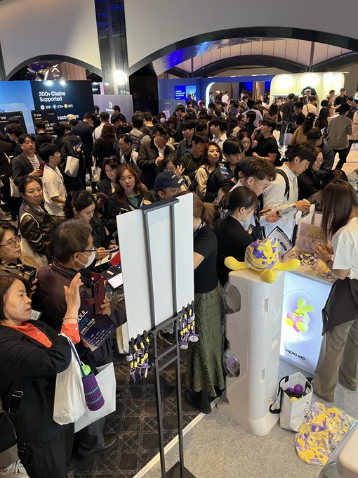 A crowded indoor convention or expo hall with attendees gathered around booths, including one displaying a 'X20' purple capsule machine and another advertising '200+ Chains Supported'.