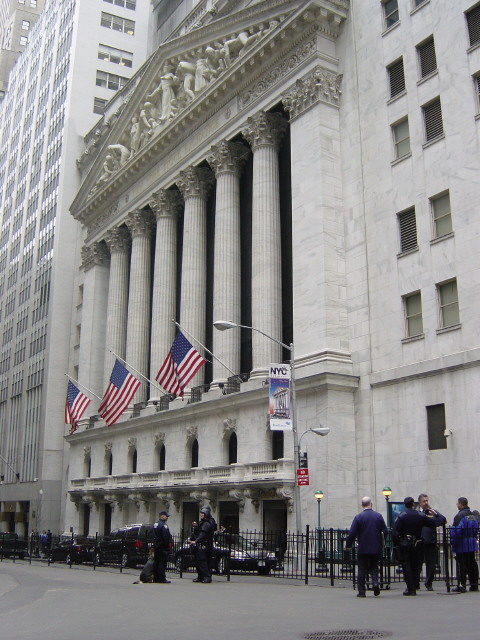 The New York Stock Exchange building is shown with its iconic columns and American flags, with police officers and pedestrians visible on the street below.