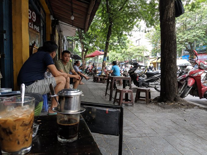 A street-level view of a Vietnamese café sidewalk scene with patrons seated at small plastic stools, motorbikes parked along the curb, and a Vietnamese iced coffee with a phin filter visible in the foreground.