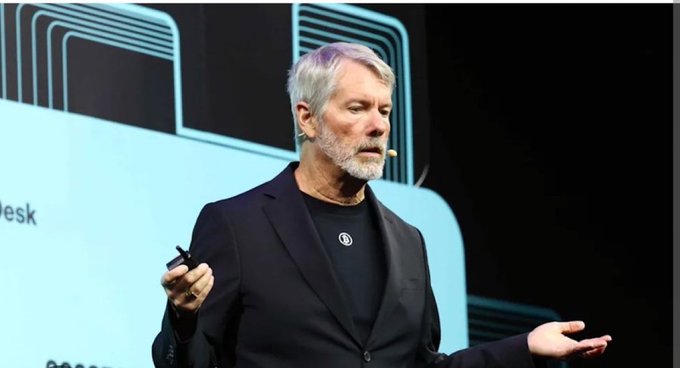 A gray-bearded man in a black blazer and t-shirt speaks on stage, holding a presentation clicker while wearing a small microphone.