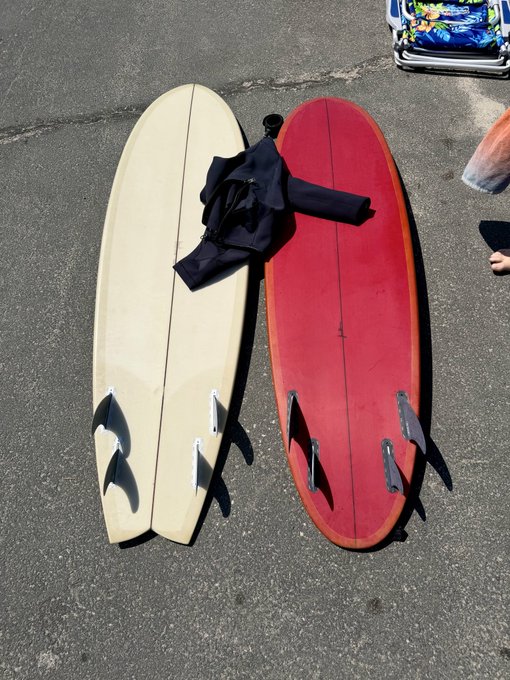 Two surfboards lying on pavement—one cream-colored and one red—both with fins visible, with a wetsuit draped on top.