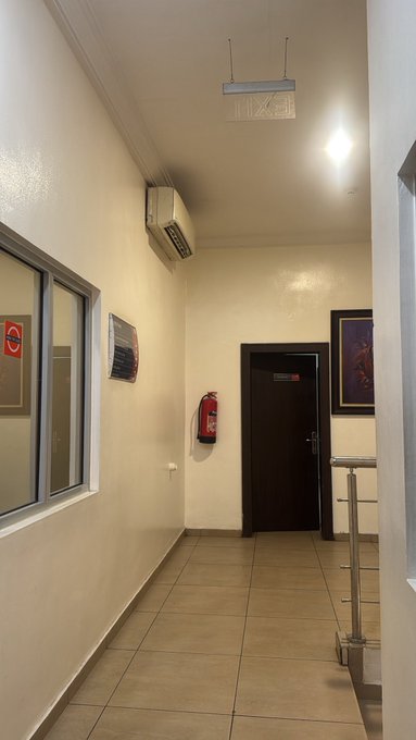 An interior hallway features white walls, tiled flooring, a wall-mounted air conditioner, a red fire extinguisher, and a dark wooden door at the end.