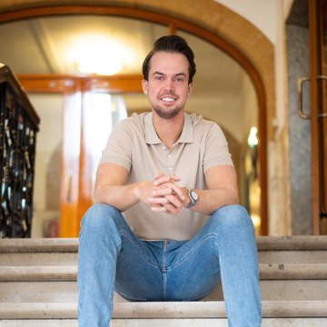 iced tea and a brown paper cup sit on the steps of an ornate wooden door, with a man in a beige shirt and blue jeans seated nearby.