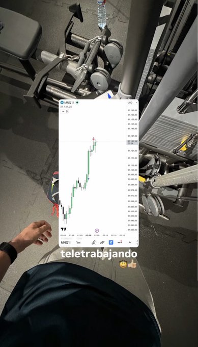 A person at the gym in shorts, sitting on a bench next to weight machines, holds a phone displaying a TradingView chart for MNQ1! with text overlay 'teletrabajando'.