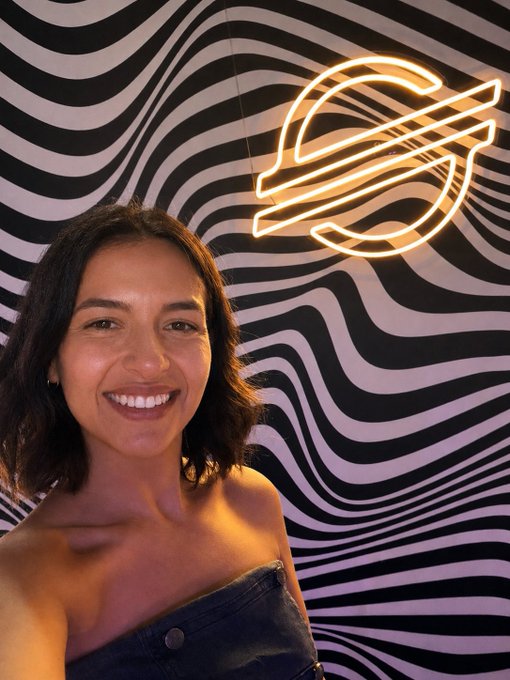 The image features a woman with short hair, wearing a black dress, standing next to a neon sign that says "S" on it. The woman appears to be posing for a photo or selfie, possibly at an event related to the "S" company. The background of the image is a black and white striped wall, which complements the neon sign. The caption in the image reads: "i spent all my energy at this amazing event now i’m having my first coffee @StellarOrg".