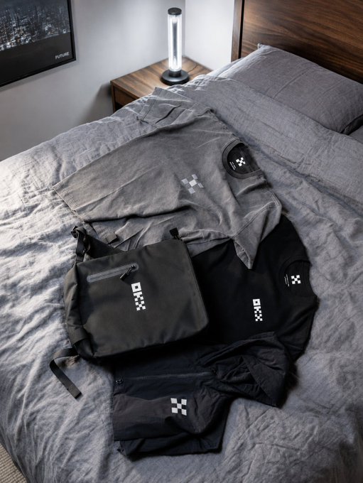 A neatly arranged bundle of grey and black OX brand clothing and a bag laid out on a grey bedsheet in a bedroom.