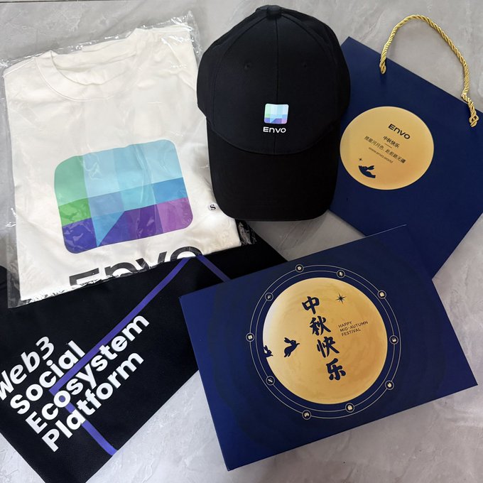 A collection of ENVO-branded promotional merchandise including a white T-shirt with a colorful logo, a black cap, a navy blue gift bag, and a Mid-Autumn Festival (中秋快乐) greeting card, alongside a 'Web3 Social Ecosystem Platform' booklet.