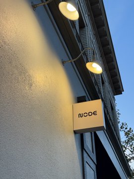 Two warm wall-mounted lights illuminate a textured white wall next to a square 'NODE' sign, set against a twilight blue sky.