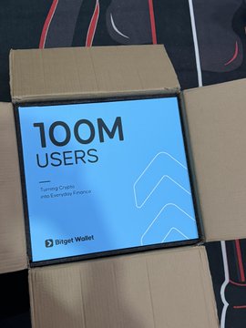 A cardboard box contains a light blue package or booklet prominently displaying "100M USERS" and "Turning Crypto into Everyday Finance" from "Bitget Wallet."
