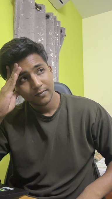 A young man in an olive green shirt looks directly at the camera while resting his head against his hand in a room with green walls.