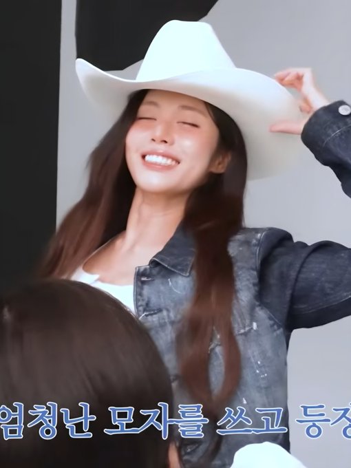 A smiling woman with long brown hair wears a white cowboy hat and a denim jacket, appearing joyful in what looks like a behind-the-scenes moment, with Korean text visible at the bottom.
