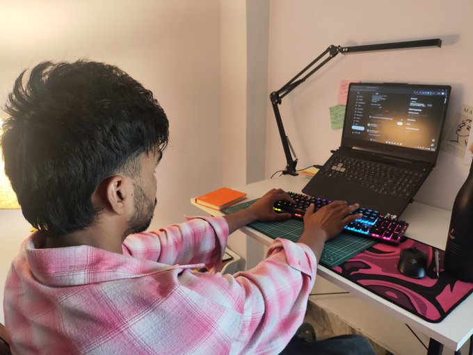 A young man in a pink plaid shirt using a mechanical keyboard with RGB lighting at his desk, with a laptop displaying code and a monitor arm lamp.