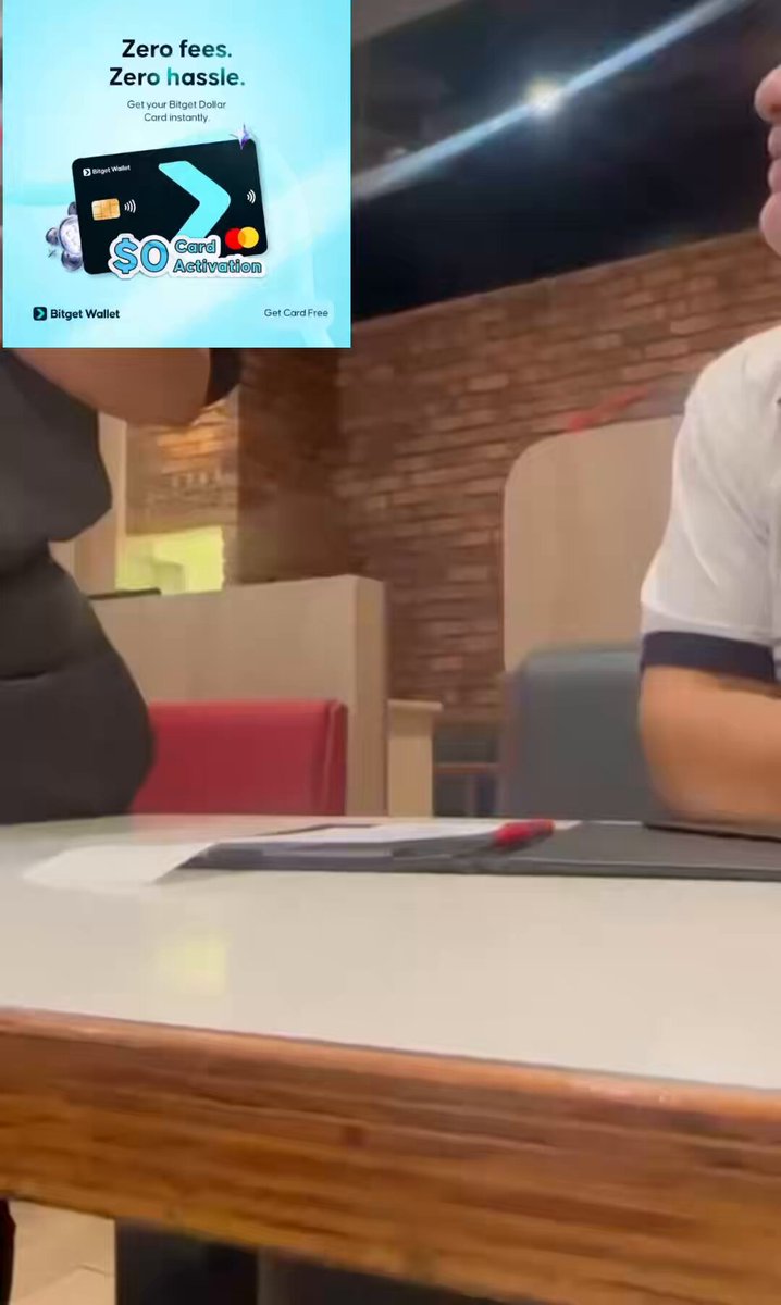 The image shows a man sitting at a table, surrounded by chairs. There are two people present - one person seated behind the other person who appears to be talking or engaged in an activity. The scene takes place in a restaurant setting with tables and chairs visible around them. A cell phone is placed on the table next to the person speaking.

The image also contains text that reads: "So guys, @BitgetWalletAF challenged us to a 20-day period where we survive using only crypto for everyday spending. Is it actually possible? Join my journey as I test it in real life. I topped up my @bitgetwallet Card with USDC on Stellar and just made my first payment."