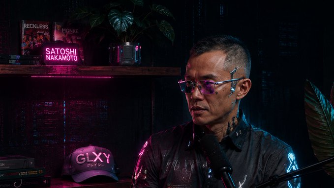 The image features a man wearing glasses and a baseball cap sitting at a desk, holding a microphone. The background includes a shelf filled with books and a plant. There are also neon signs visible in the scene. The image has a caption that reads "CZ stands his ground. I wonder if this is an attempt to save the minds of the remaining people who have completely lost faith in the market, or just talk for the sake of talking?".