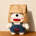 A small plush figure wearing a brown paper bag over its head sits on a wooden surface, dressed in blue denim overalls.
